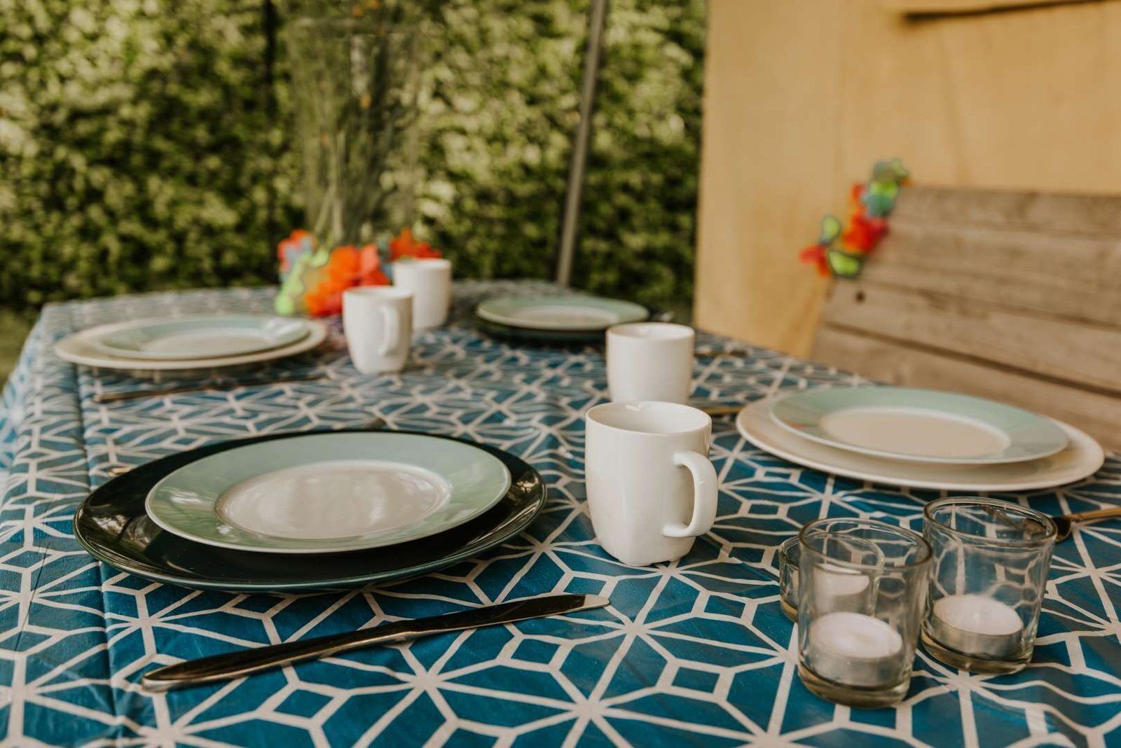 Un tavolo apparecchiato con piatti, una tazza, posate e bicchieri, con una tovaglia decorata e una parete di legno sullo sfondo.