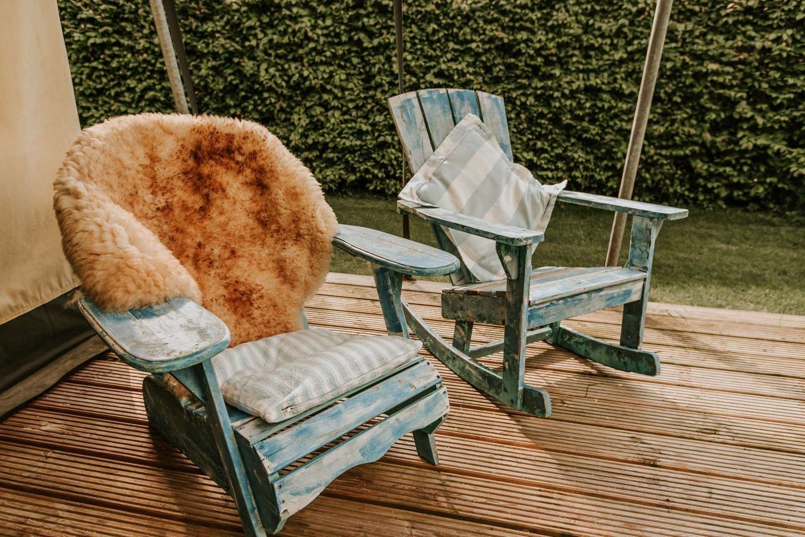 Deux chaises en bois rustiques, dotées de coussins, dont l’une a un siège recouvert de fourrure, sont placées sur une terrasse en bois, avec une haie en arrière-plan.