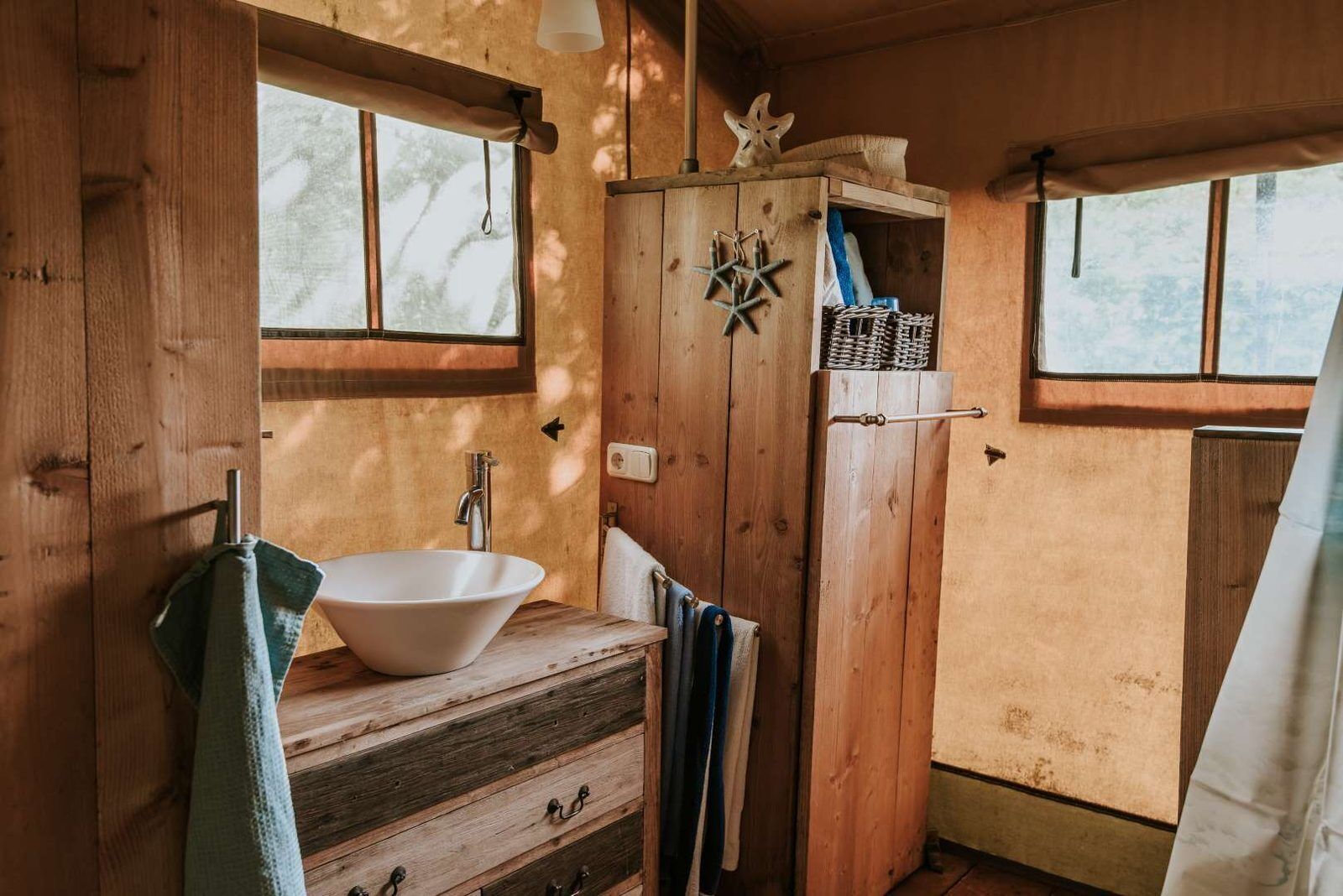Een houten badkamer in een hutje, met een groot raam, een houten kastje en een witte wastafel.