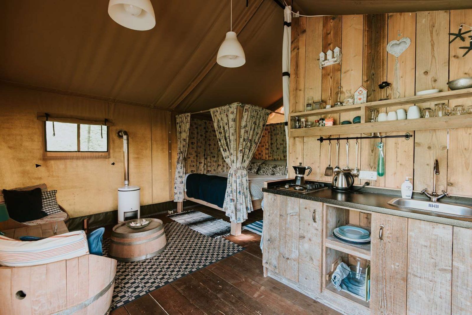 Ein rustikales Blockhaus-Interieur mit einem Holzesstisch, einem Bett mit gemusterten Vorhängen, einer Küchenzeile mit Holzschränken und einem Spülbecken sowie einem großen Fenster.