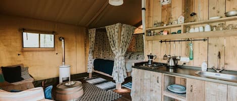Interior rústico de cabana com mesa de jantar de madeira, cama com cortinas estampadas, área de cozinha com armários de madeira e pia, e uma janela grande.