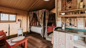 A rustic wooden room with a four-poster bed, a small table, and a shelf with kitchen items. - Kampari - Luxury Safari Tent - Out of Africa (Delfstrahuizen)