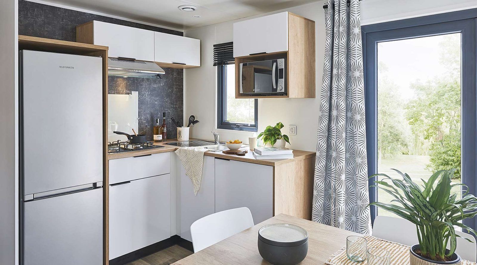 Une cuisine moderne avec des meubles blancs, un four à micro-ondes encastré et une fenêtre agrémentée de rideaux à motifs.