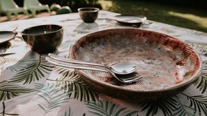 A table with a leaf-patterned tablecloth, a metal bowl, a spoon, and a fork. - Kampari - Tiny House - Bosk (Delfstrahuizen)