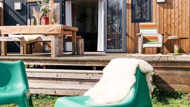 A wooden deck with a table and chairs, a sliding glass door, and a small outdoor seating area. - Kampari - Tiny House - Bosk (Delfstrahuizen)