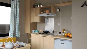 Eine kompakte Küche mit Spüle, Herd und kleinem Kühlschrank. Über der Arbeitsfläche befinden sich Schränke und ein Fenster mit gemusterten Vorhängen.