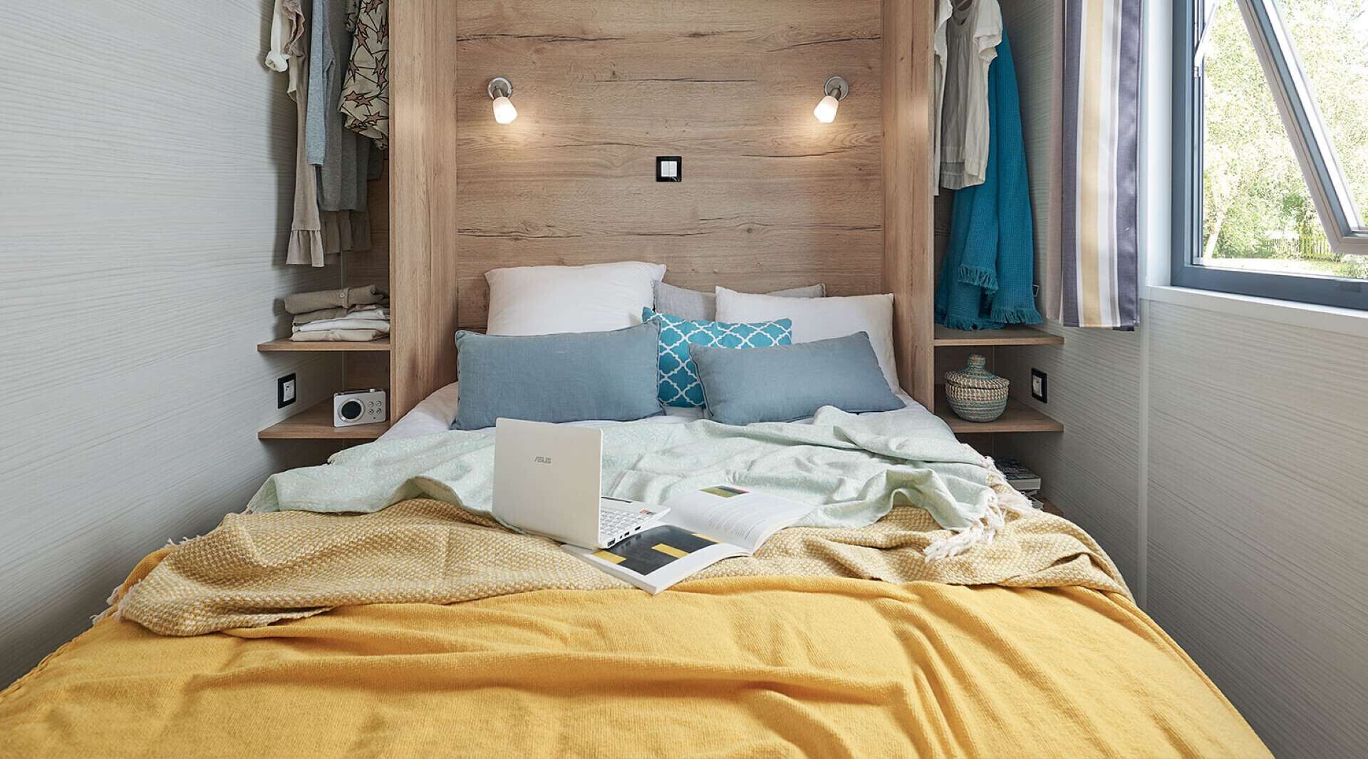 Un lit avec un ordinateur portable, des oreillers et une couverture. Il y a des étagères avec des vêtements et une radio.