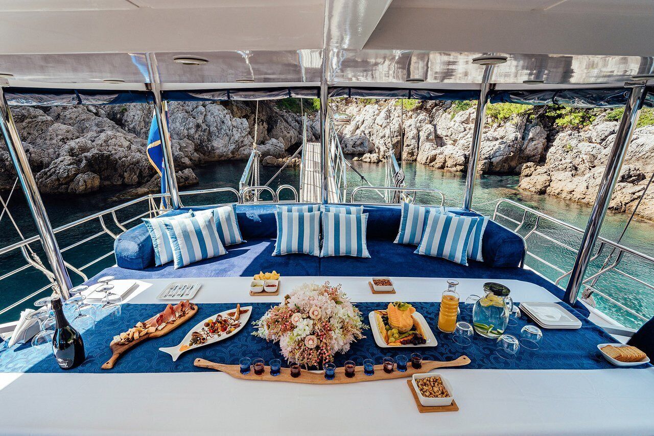 A boat deck with a dining table set for a meal, surrounded by blue cushioned seating.