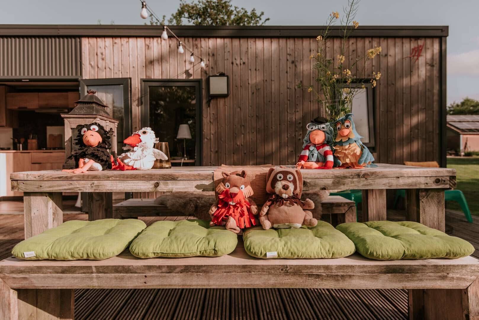 A wooden table with plush toys and green cushions in front of a wooden building.