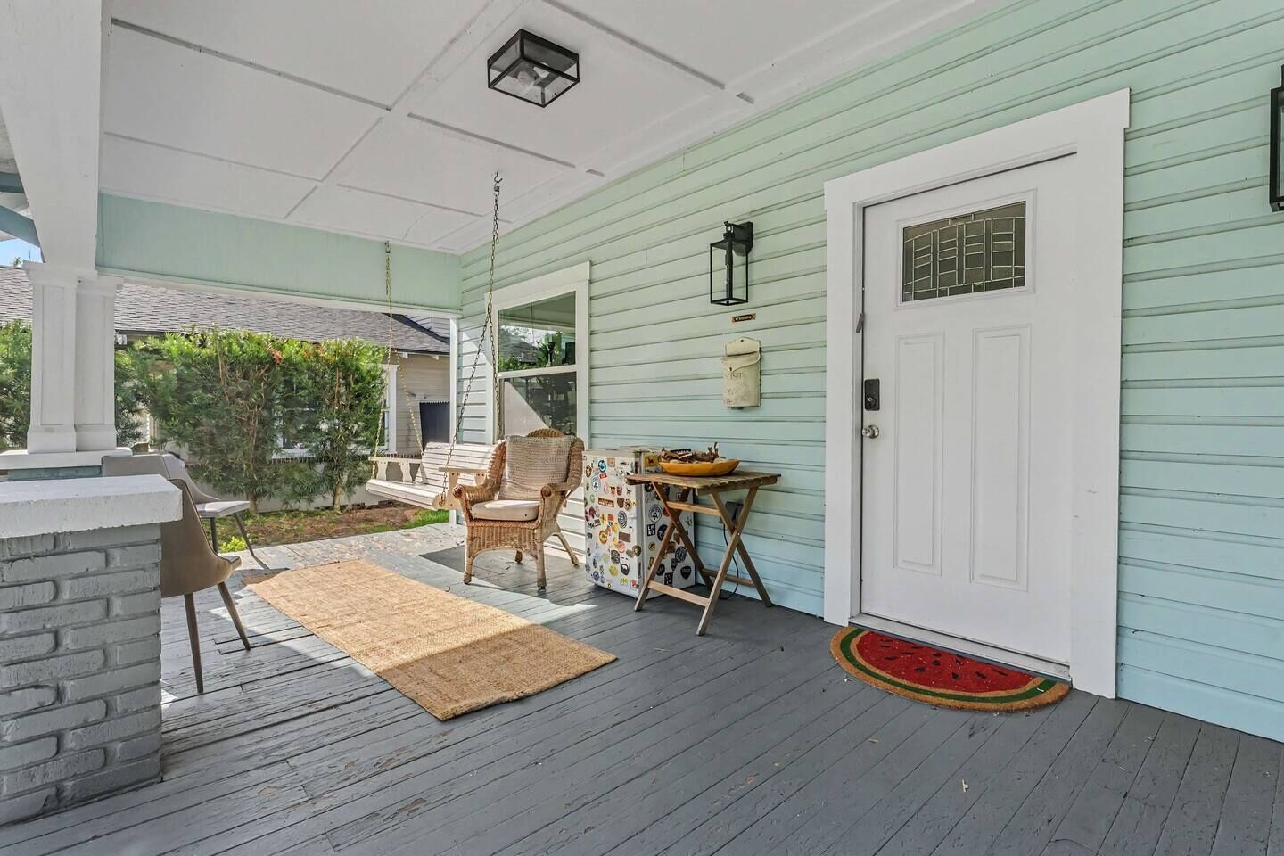 A covered porch with a wicker chair, a small table with a bowl, and a door with a window.