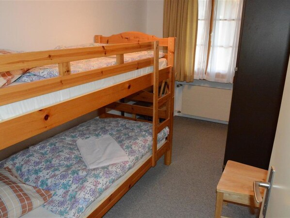A bunk bed with a wooden frame, a small wooden stool, and a window with curtains.