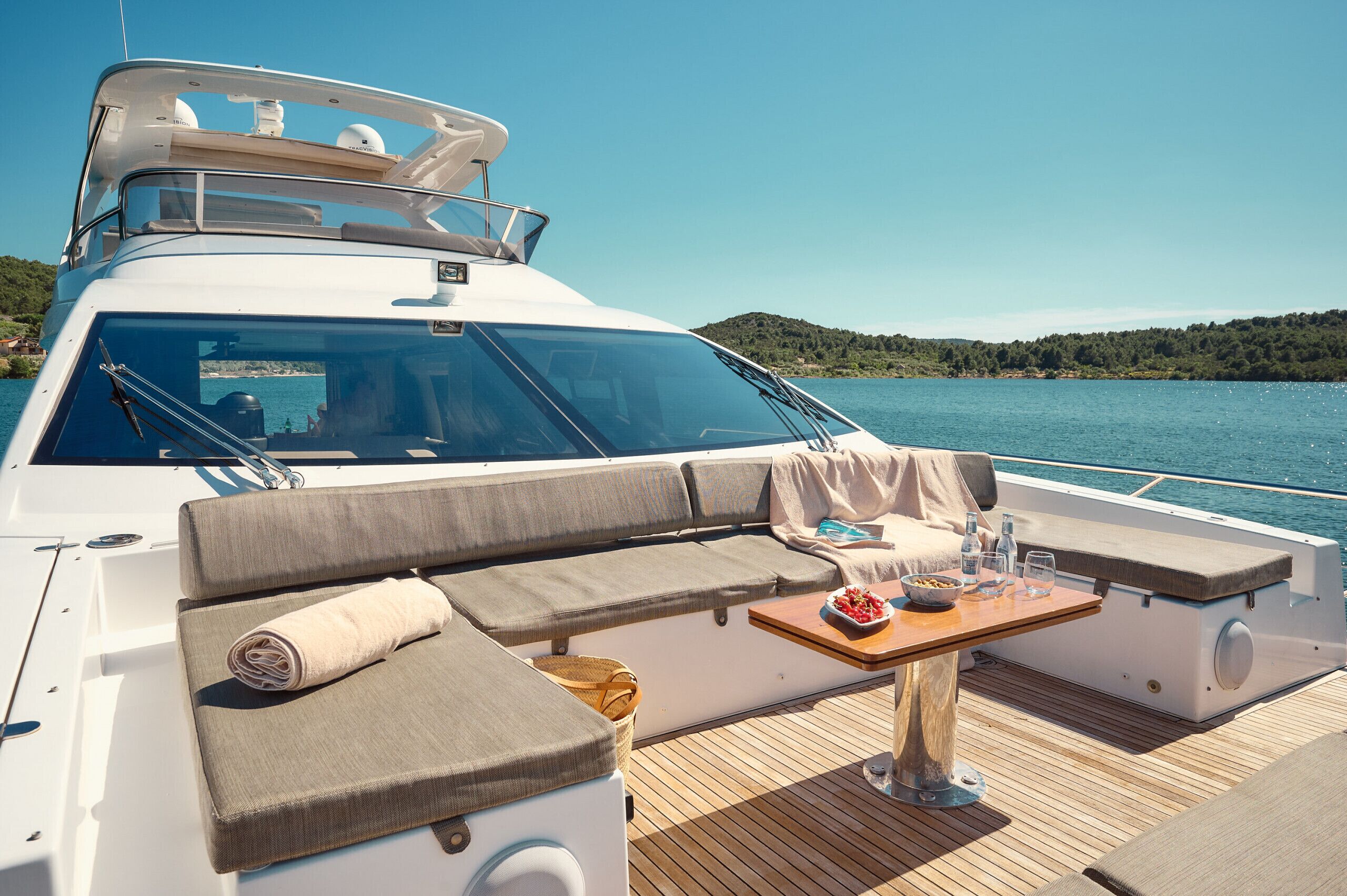 A yacht with a covered seating area, a table with drinks, and a view of the water and hills.