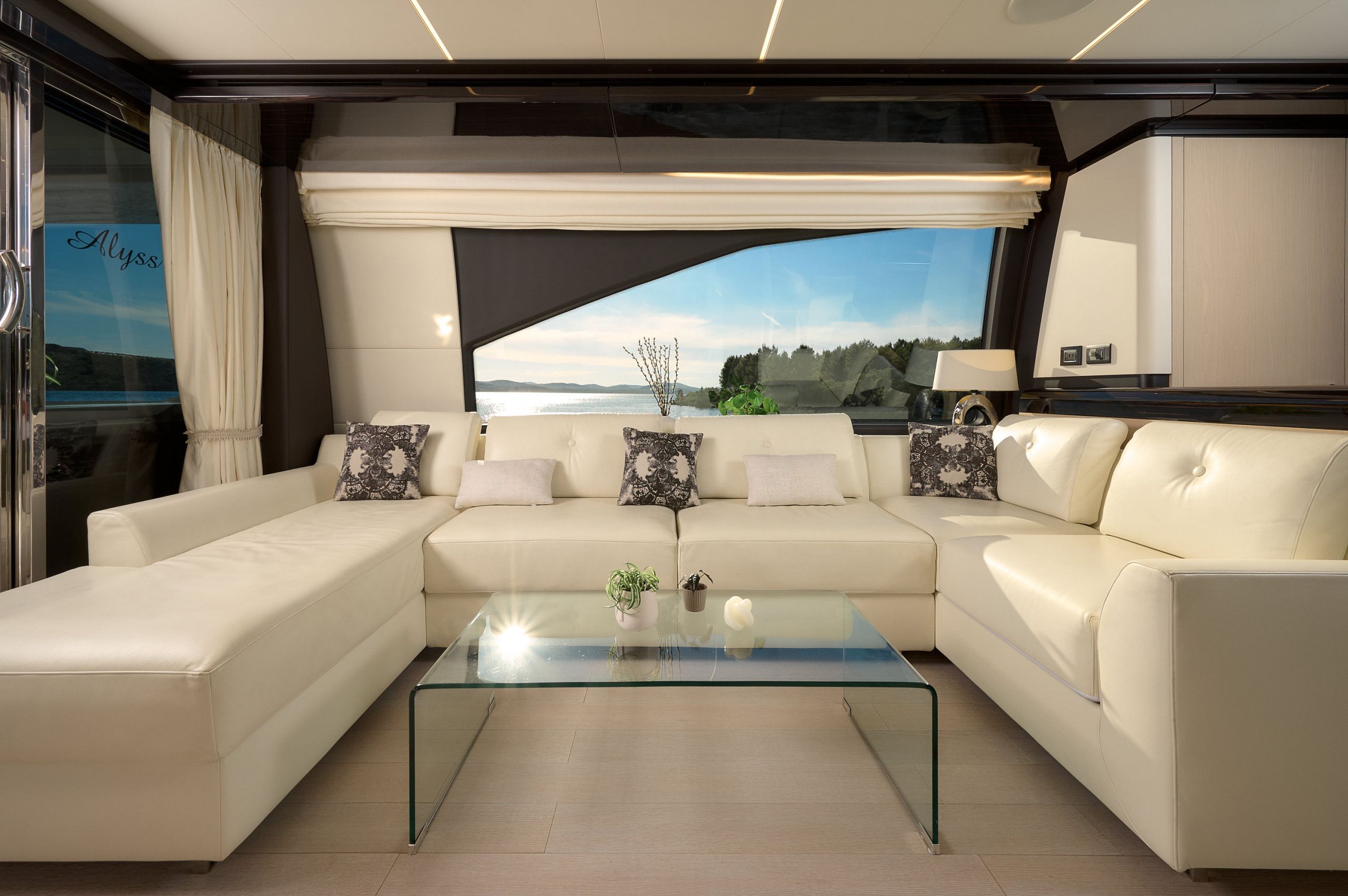 A modern living area with a glass coffee table, two white sofas with patterned cushions, and a large window offering a scenic view.