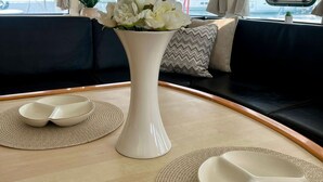 L'intérieur d'un bateau avec une table à manger, deux assiettes blanches, un vase avec des fleurs blanches et une vue sur le port de plaisance à travers les fenêtres.