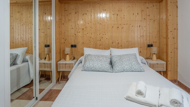 A bedroom with a wooden paneled wall, a bed with white bedding, two bedside tables with lamps, and a mirror on one wall.