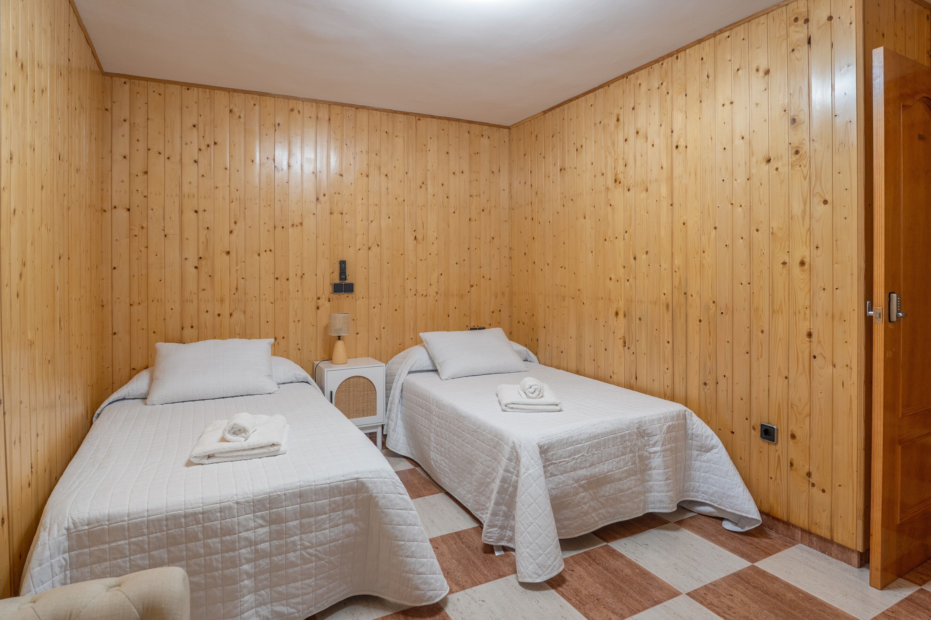 Una habitación con dos camas, una mesita de noche, una lámpara y una puerta de madera.