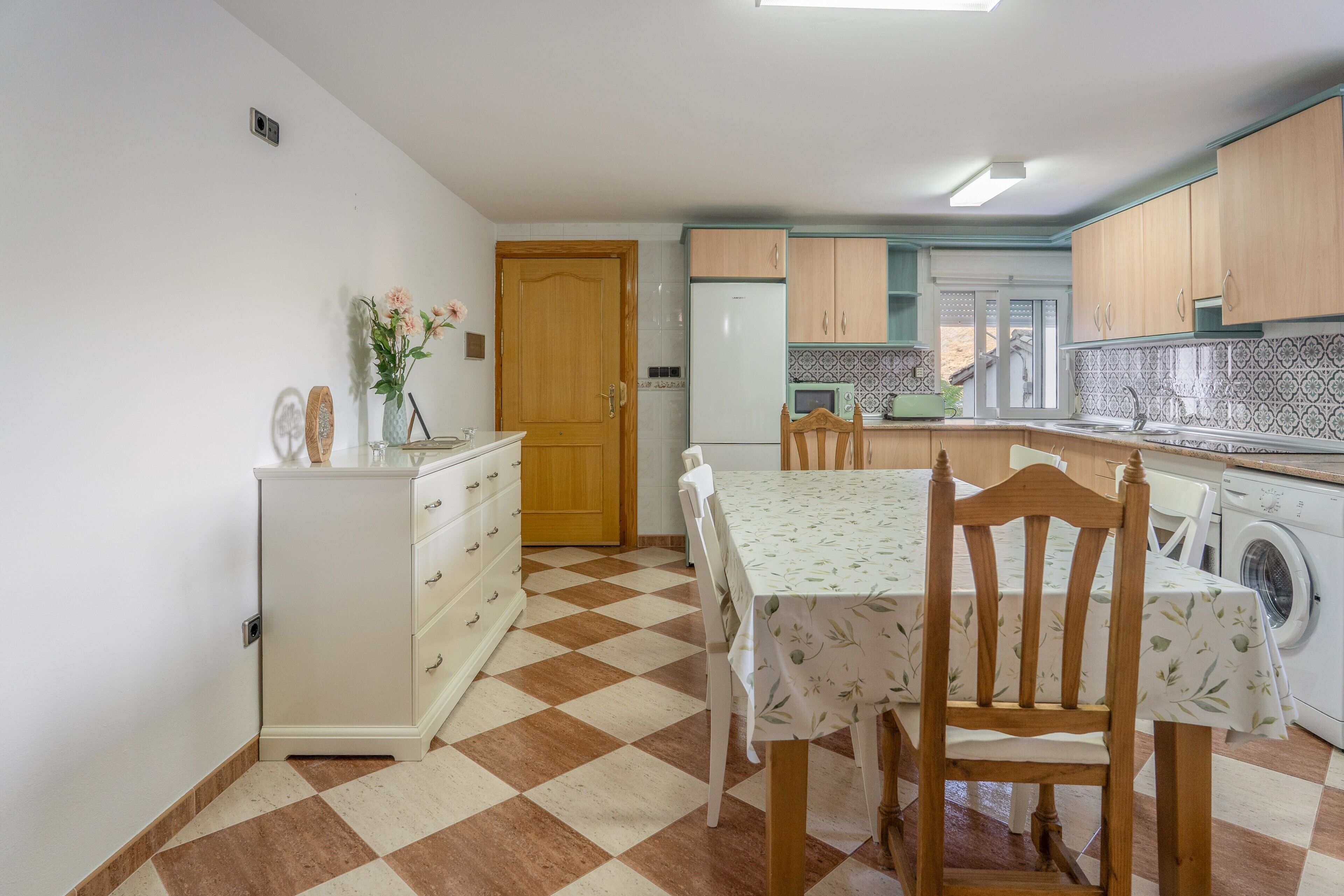 Una cocina con piso a cuadros, armarios de madera, un refrigerador, una lavadora y una mesa de comedor con sillas.