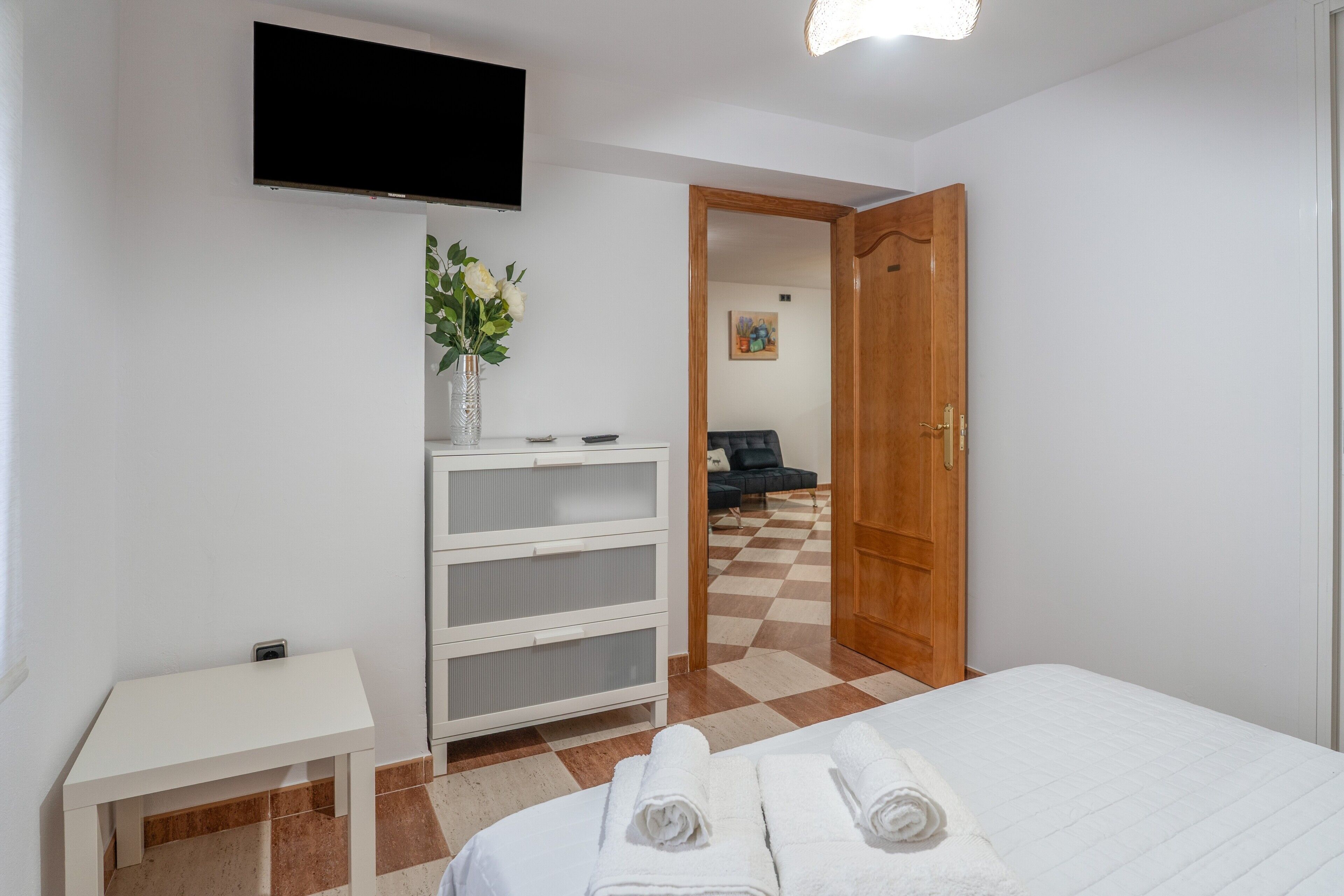 Une chambre d’hôtel avec un lit, un téléviseur fixé au mur, une petite table ornée d’un vase de fleurs et un sol à carreaux.