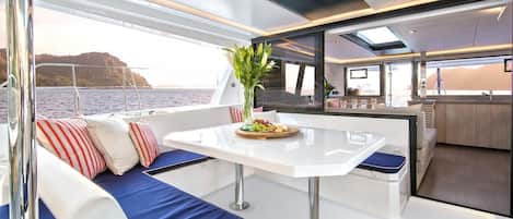 A modern boat interior with a dining area, seating with cushions, a table with a fruit platter, and a view of the water.