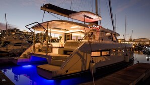 Un yacht était amarré dans une marina, avec un pont bien éclairé, un espace salon couvert et une plateforme flottante dotée d’escaliers menant au pont supérieur.