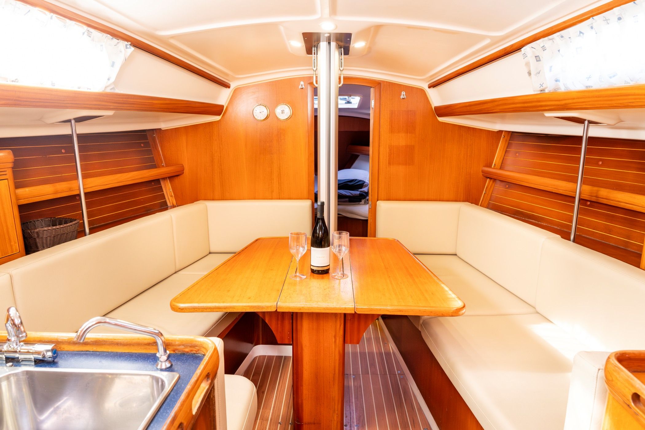 A boat interior with a wooden table, cushioned seating, a sink, and a window with blinds.