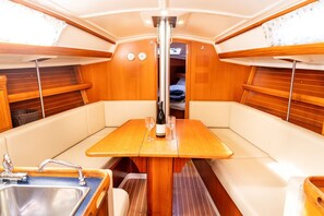 A boat interior with a wooden table, cushioned seating, a sink, and a window with blinds.