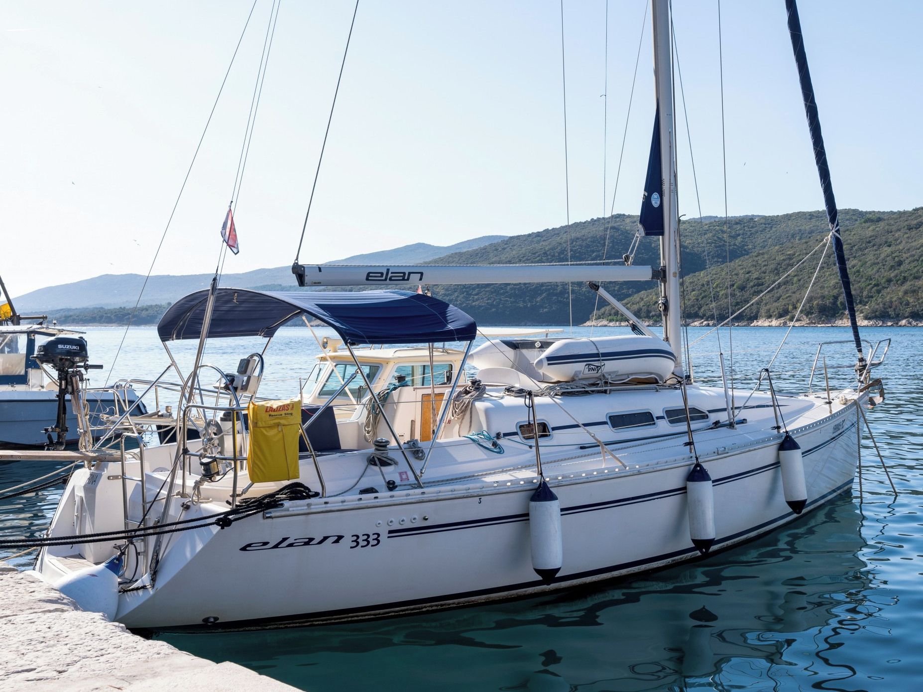 A sailboat named 'elan 333' docked at a marina with clear water and a hilly landscape in the background.