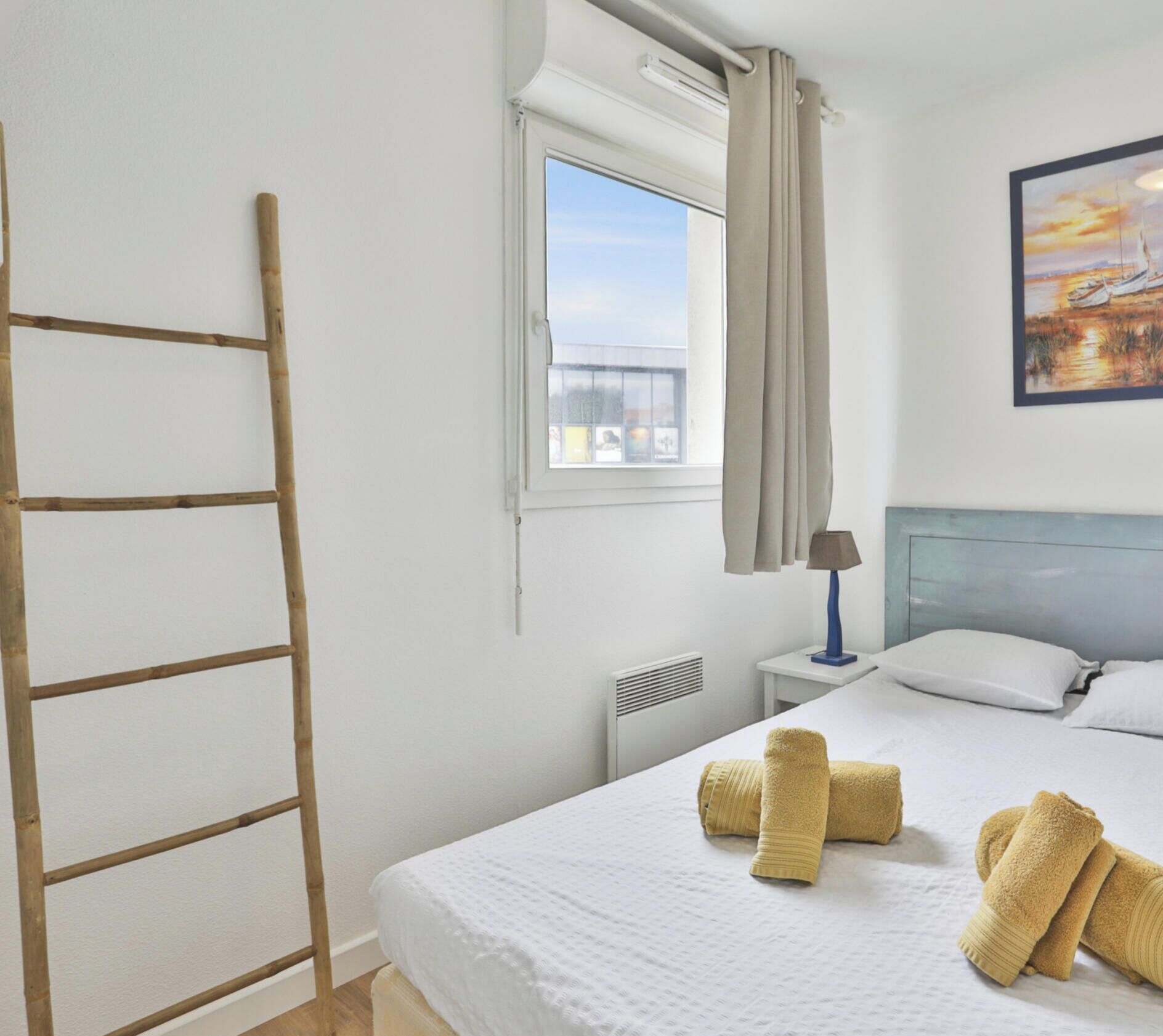 Una habitación de hotel con cama, una ventana con cortinas, una mesita de noche con lámpara y un cuadro en la pared.