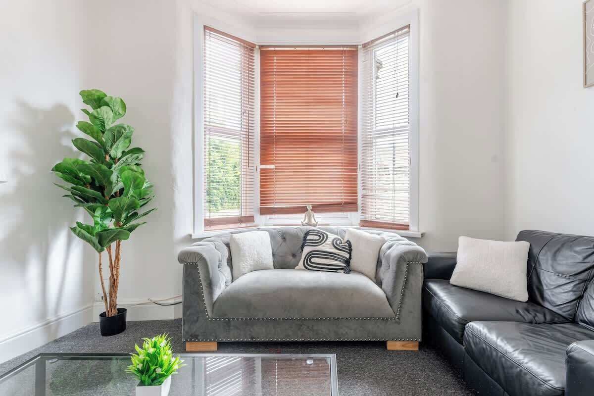 A living room with a grey sofa, a black leather sofa, a potted plant, and a bay window with wooden blinds.