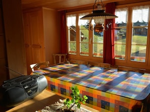 Ein Zimmer mit einem großen Tisch, der mit einer farbenfrohen Steude bedeckt ist, Holzvertäfelung, einem Radio und einem Fenster mit Vorhängen.