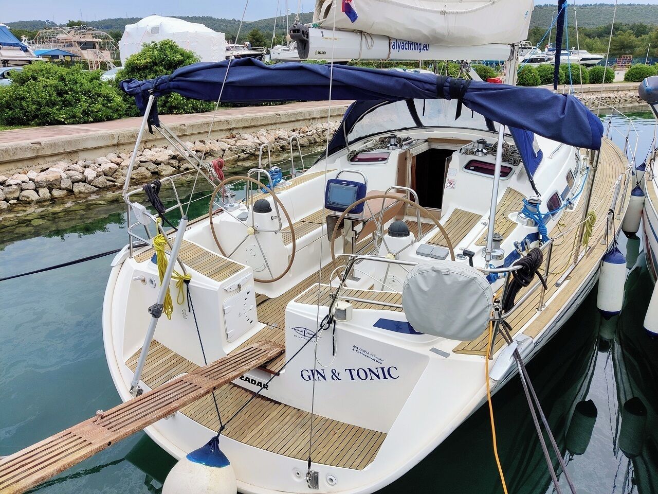 Een zeilboot afgemeerd in een jachthaven met een overdekt dek, een stuurwiel en de naam 'GIN & TONIC' op de boeg.