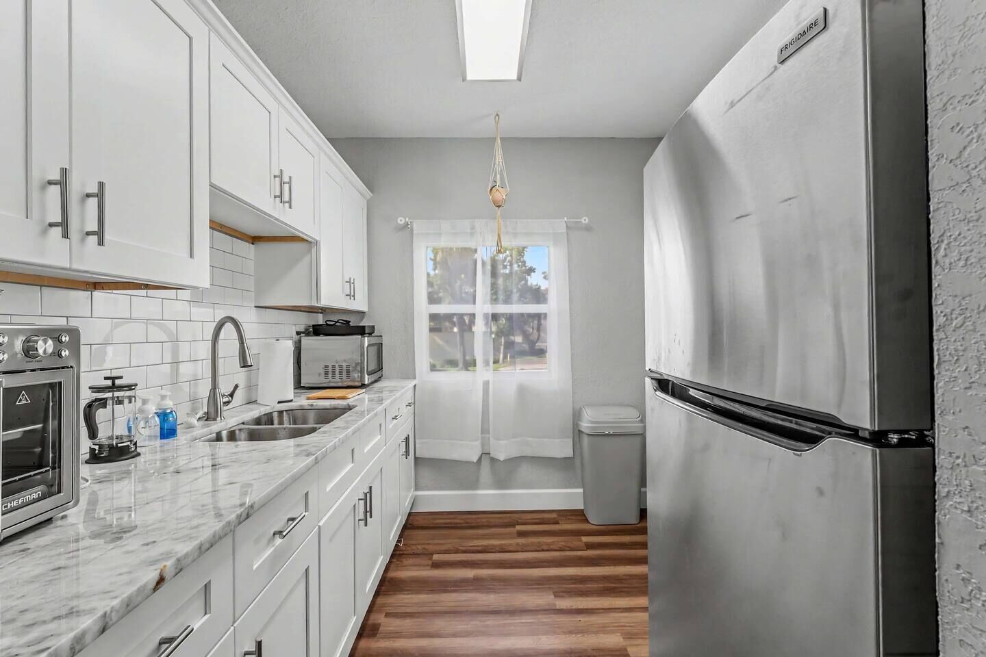 A compact kitchen with white cabinets, a stainless steel refrigerator, a built-in oven, a microwave, and a window with a curtain.