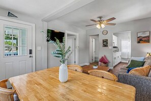Un coin repas avec une table en bois, des chaises, un vase avec des plantes, un ventilateur de plafond et, au fond, une cuisine.
