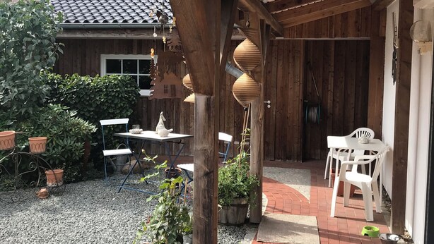 Un patio couvert avec un sol en gravier, des meubles en bois, des plantes en pot et une maison en arrière-plan.