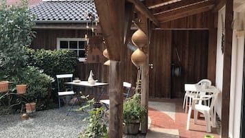 Eine überdachte Terrasse mit Kiesboden, Holzmöbeln, Topfpflanzen und einem Haus im Hintergrund.