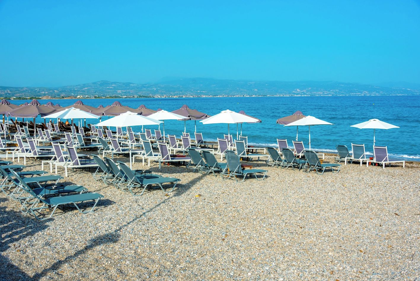 Une plage bordée de rangées de chaises longues et de parasols, une plage de galets, et la mer en arrière-plan.