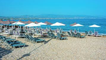 Une plage bordée de rangées de chaises longues et de parasols, une plage de galets, et la mer en arrière-plan.