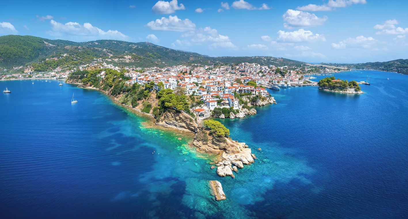 An aerial view of a coastal town with buildings, clear blue waters, and a hilly landscape in the background.