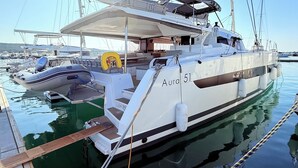 En hvid sejlbåd, der hedder 'Aura 51', ligger til anker i en havn blandt andre både, med en overdækket dæk og et wakeboard.