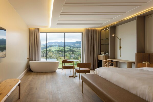 A modern hotel room with a large bed, a sitting area with a table and chairs, a bathtub, and a view of a green landscape.