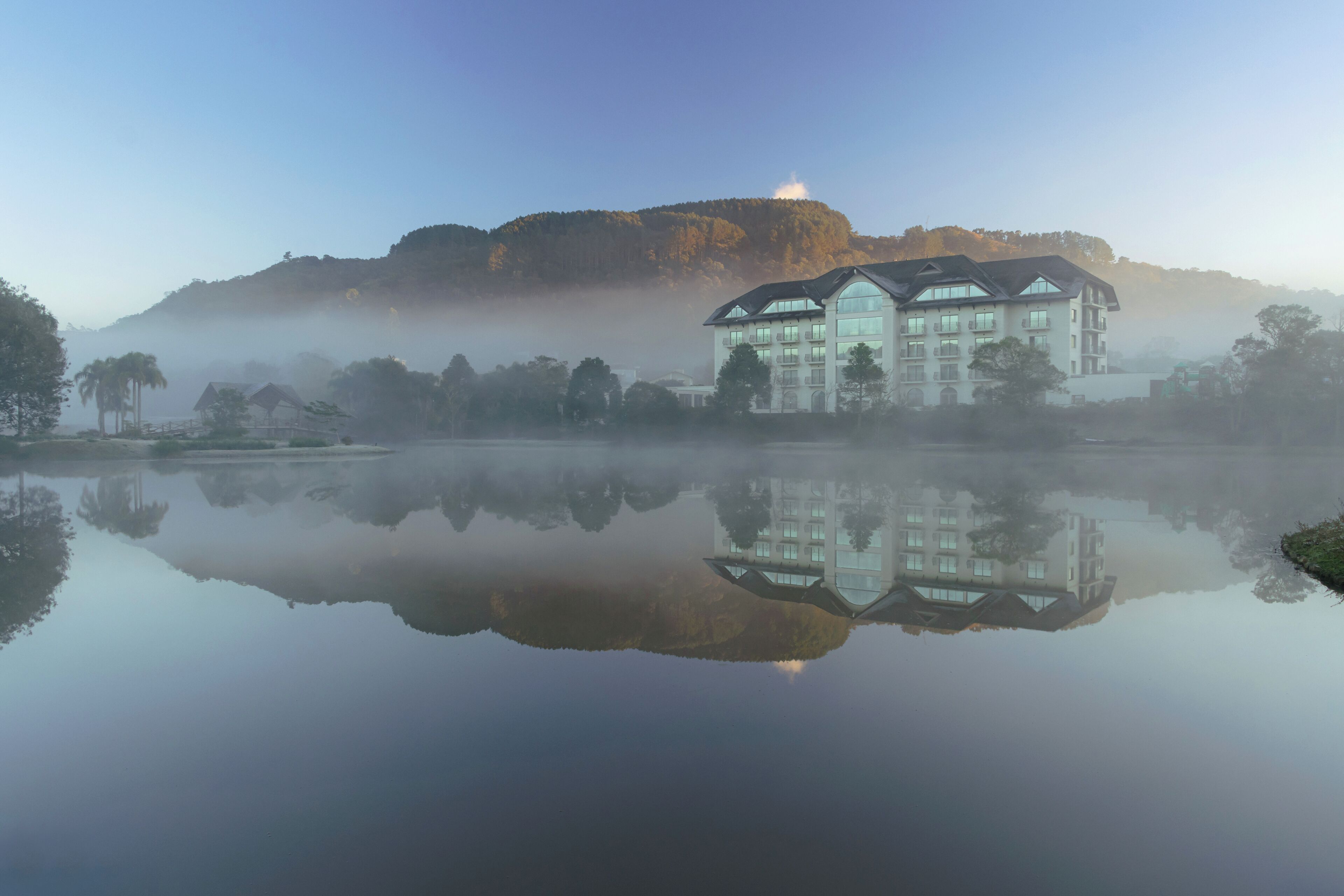 A large building with multiple floors and a flat roof, situated near a calm body of water with a reflection.