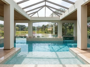 A swimming pool with a glass enclosure, surrounded by a landscaped garden and a view of a lake.