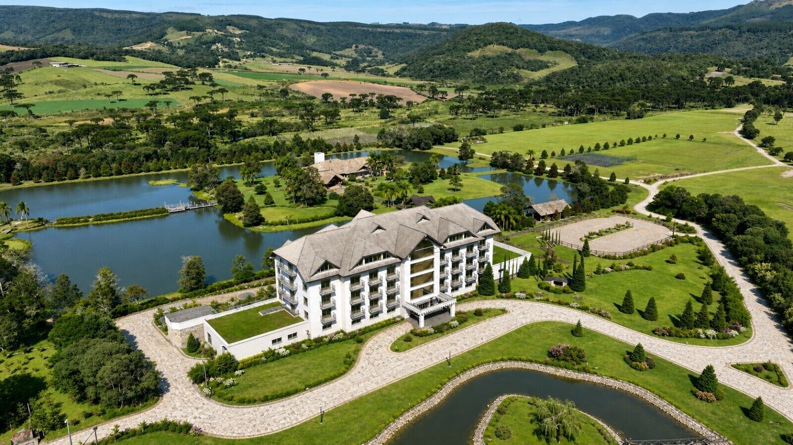 Un hotel grande de varios pisos con un patio central, rodeado de jardines ajardinados y un lago.