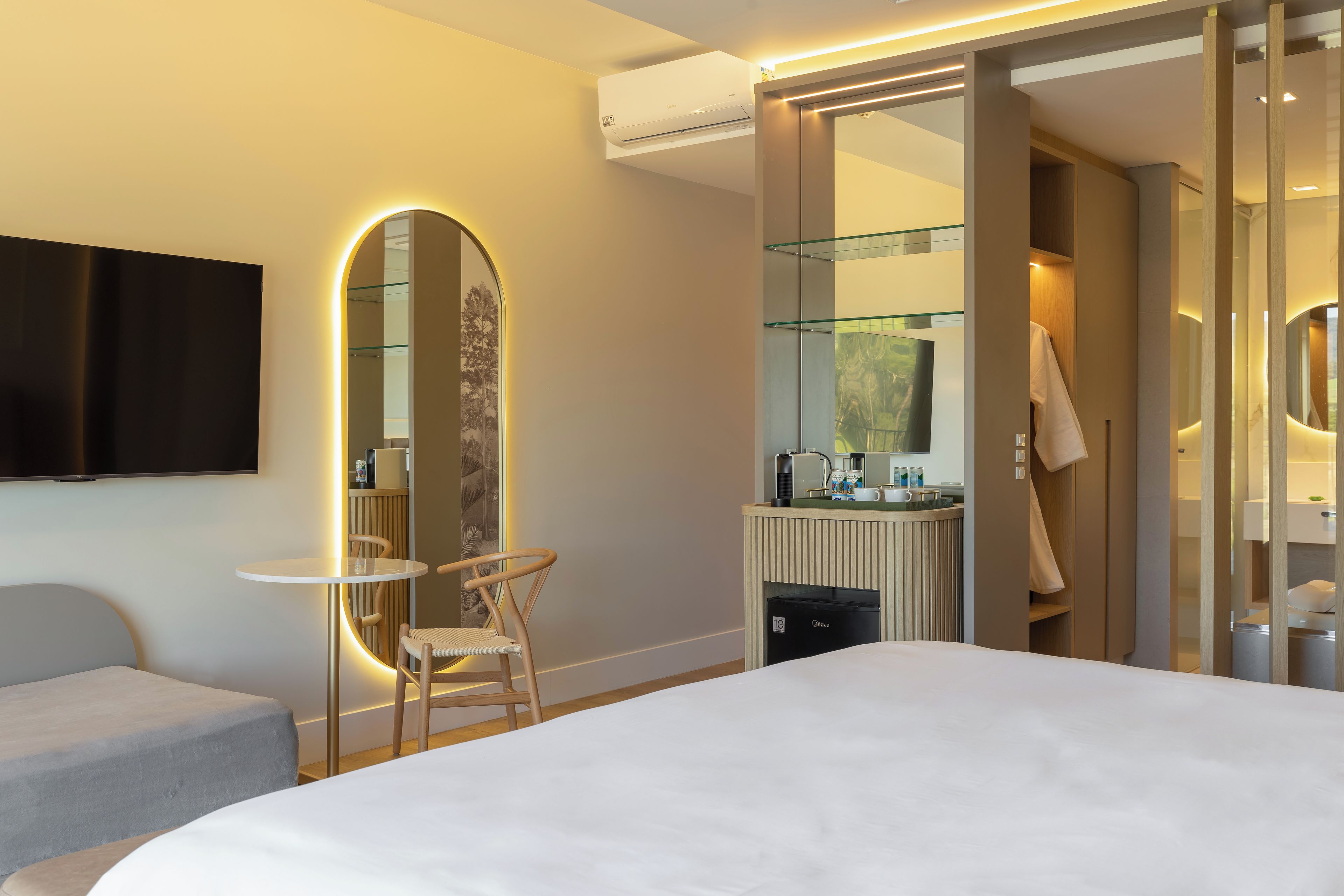 A modern hotel room with a large bed, a small table with a chair, a large mirror, a television, and a bathroom with a glass shower enclosure.