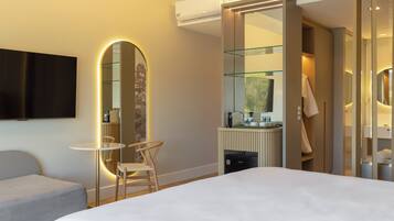 A modern hotel room with a large bed, a small table with a chair, a large mirror, a television, and a bathroom with a glass shower enclosure.
