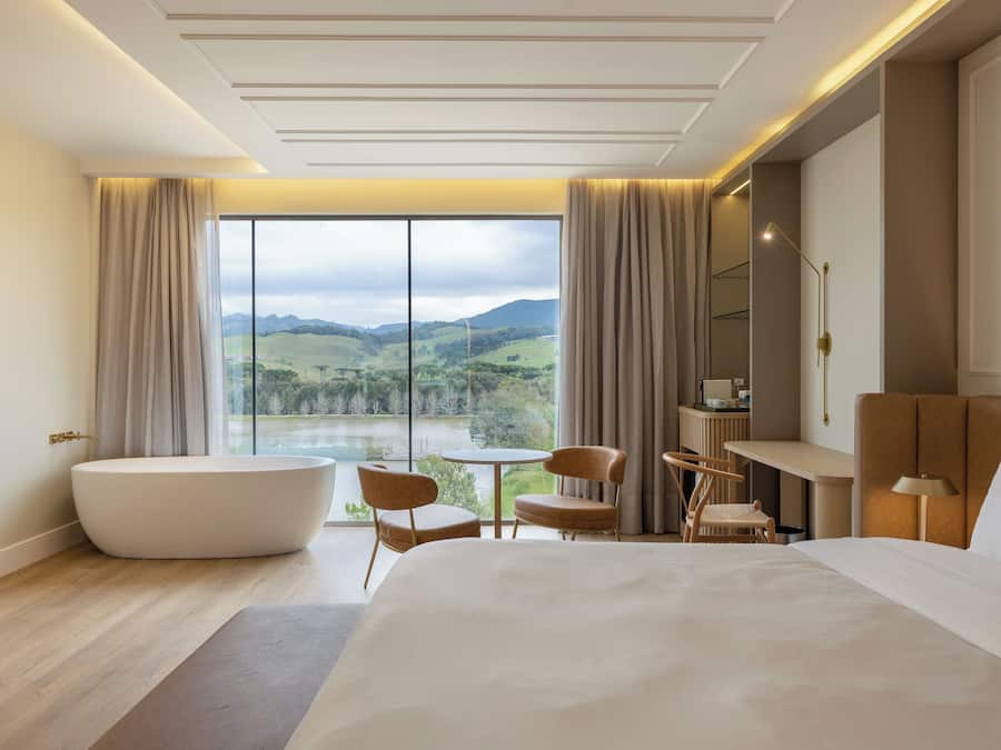 A modern hotel room with a large bed, a bathtub, a dining table, and a view of a lake and mountains.