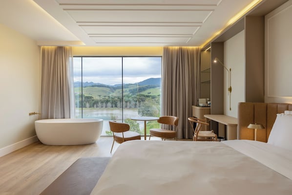 A modern hotel room with a large bed, a bathtub, a dining table, and a view of a lake and mountains.