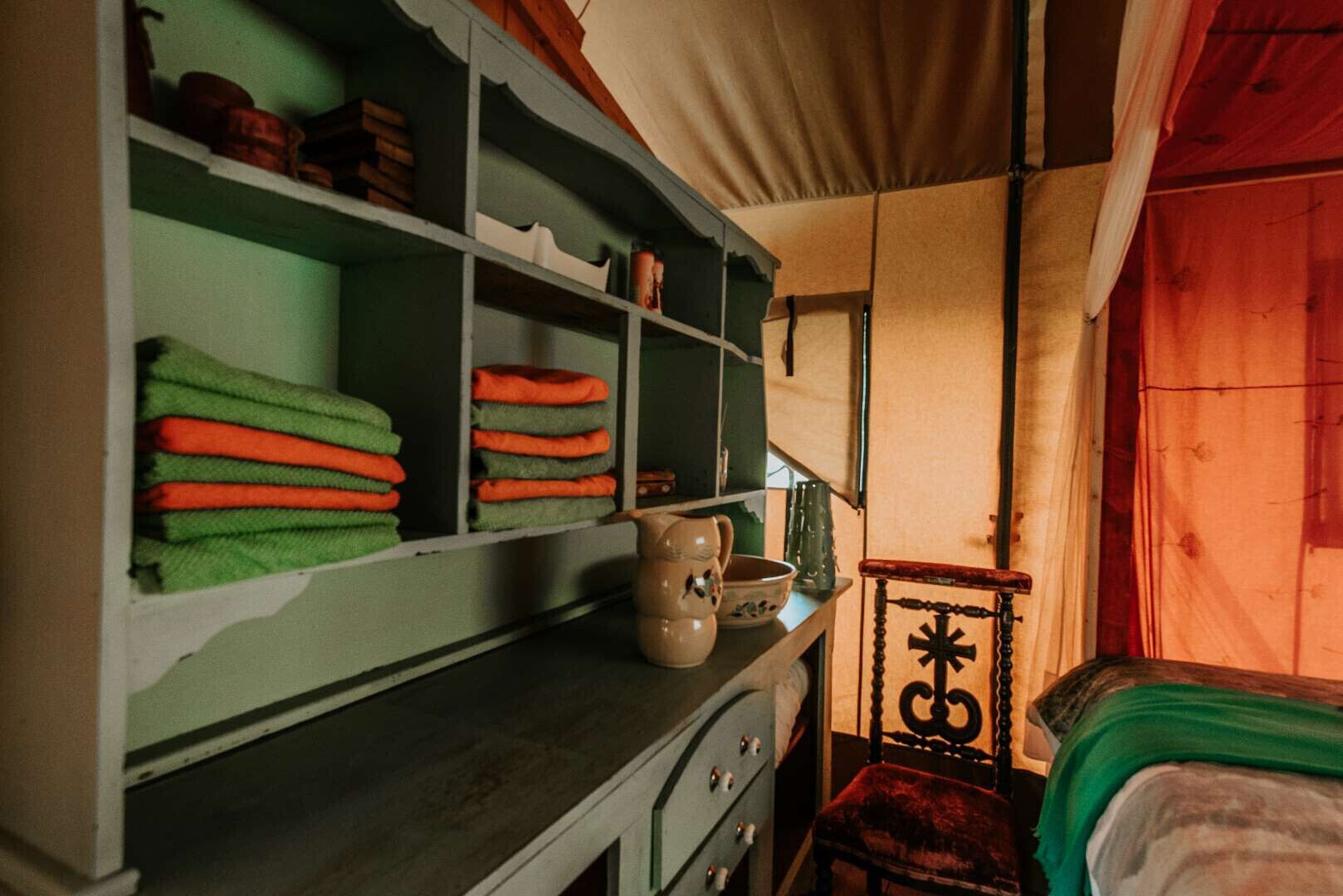Une pièce comprenant un lit, une étagère avec des serviettes pliées, une chaise en bois et une table sur laquelle se trouvent une carafe et un bol.