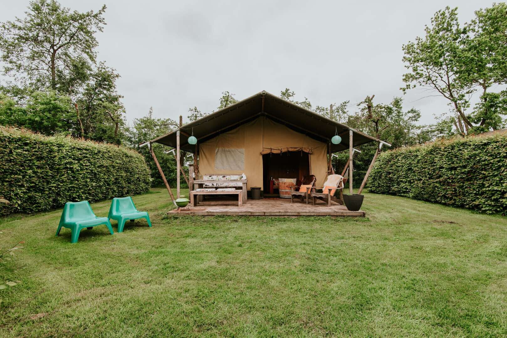 Acomodação ao ar livre em tenda, com deck de madeira, duas cadeiras de plástico verde e uma mesa.