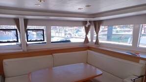 Uma cabana com mesa e bancos, janelas amplas com cortinas e vista para o porto de embarcações.